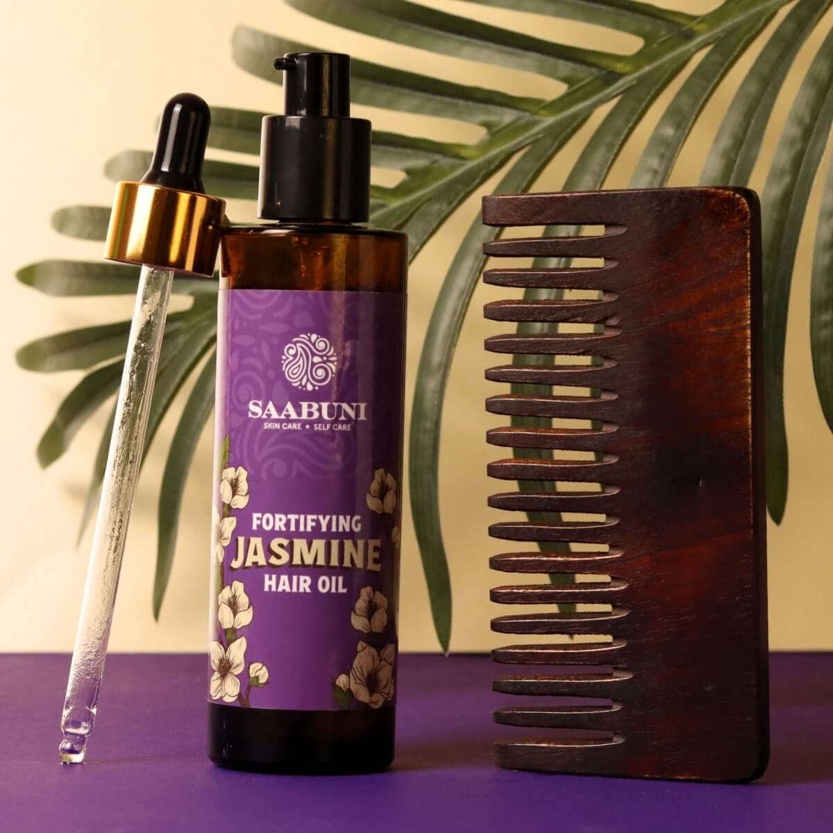 Saabuni Jasmine hair oil with a dropper and wooden comb on a purple surface with green leaves in the background