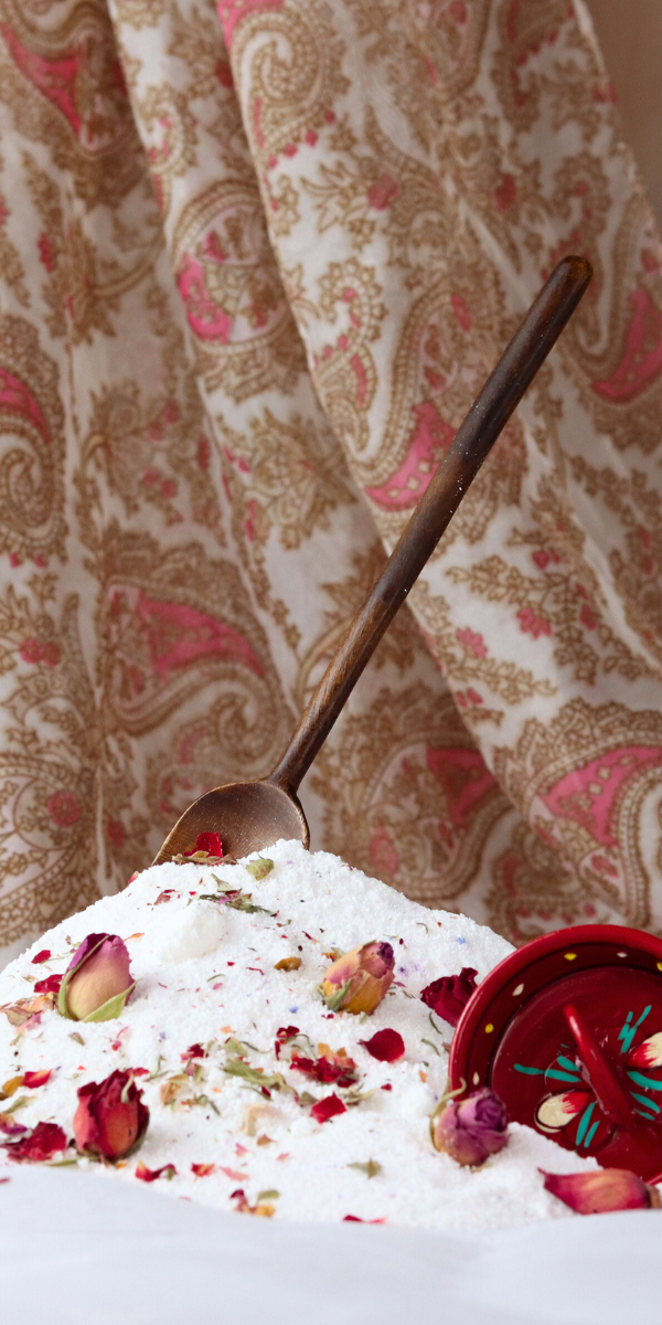Decorative bath salts with rose petals and a spoon on a textured surface