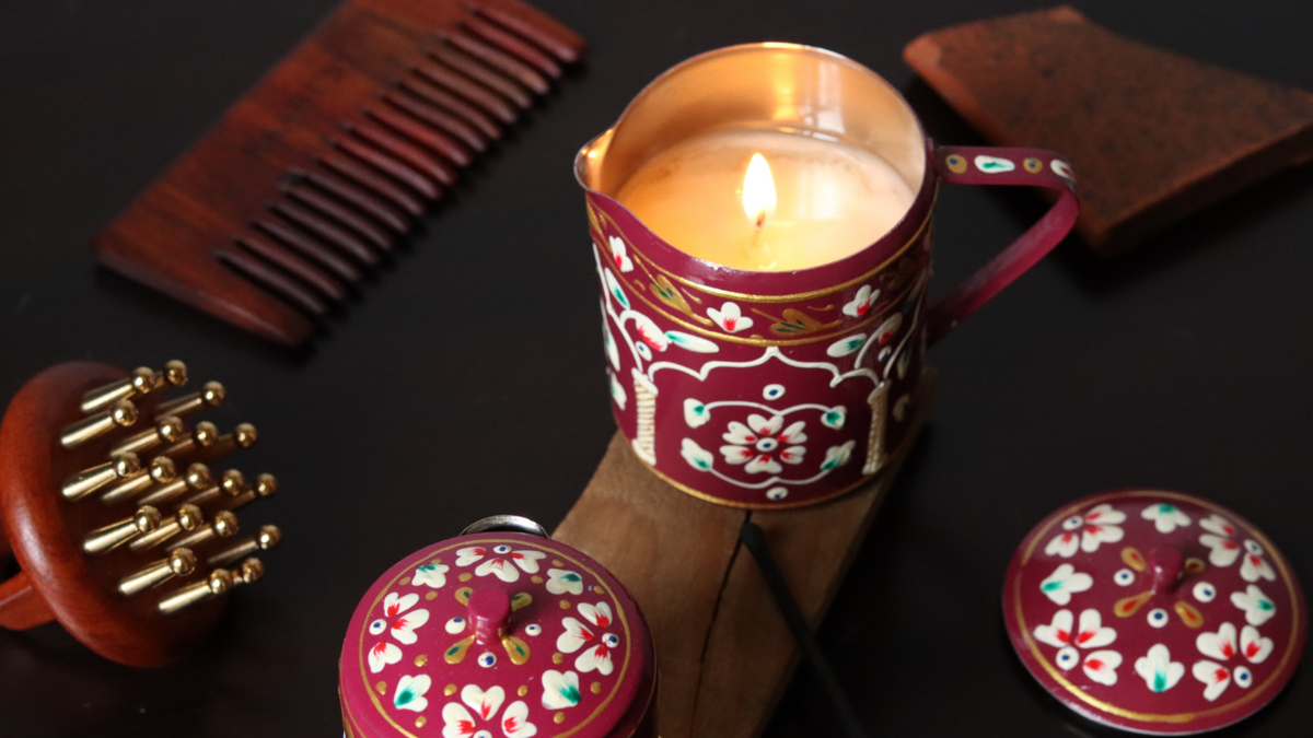 A decorative red metal candle holder with floral patterns and a lit candle, styled on a dark surface with wooden hair tools and accessories around it.