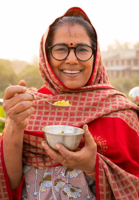 Woman in a red and pink checkered scarf holding a bowl and spoon outdoors.