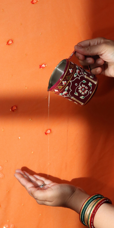A close-up of hands holding a small ornate metal cup with a floral pattern, pouring a thin stream of oil onto an open palm against a bright orange background with scattered red flower petals.