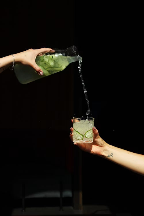 Person pouring a green liquid from a bottle into a glass against a black background